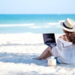 A woman using and typing on laptop computer keyboard while sitting on a beautiful beach