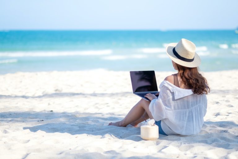 A woman using and typing on laptop computer keyboard while sitting on a beautiful beach